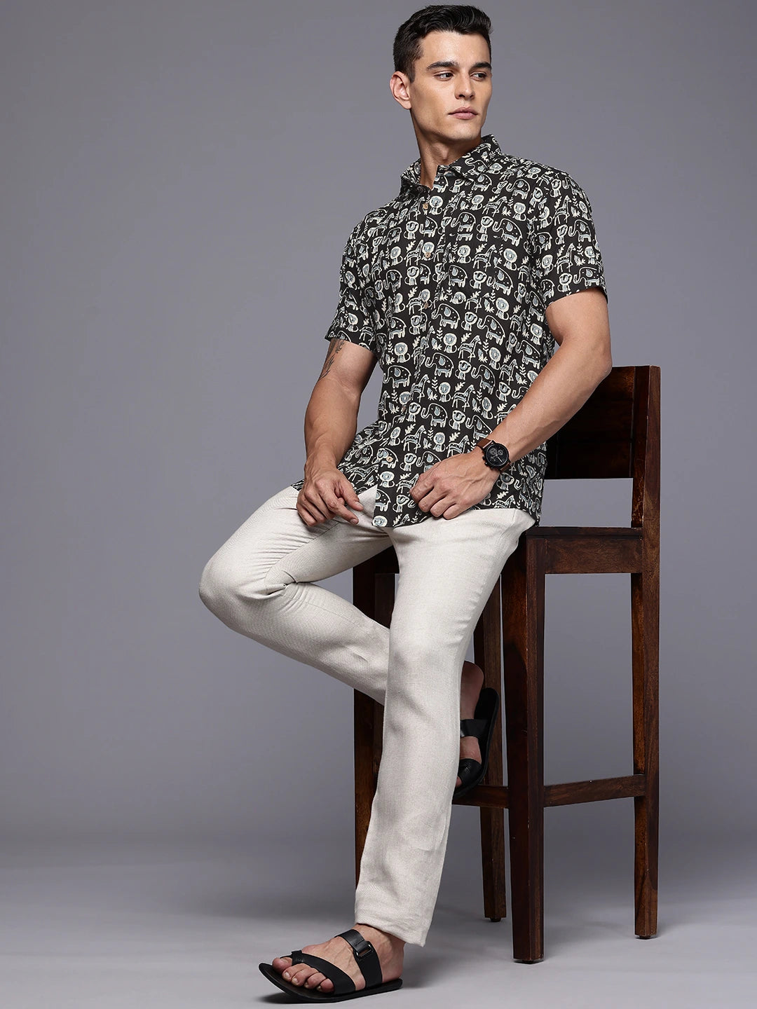 Man sitting on a wooden stool wearing a printed shirt and white pants against a gray background