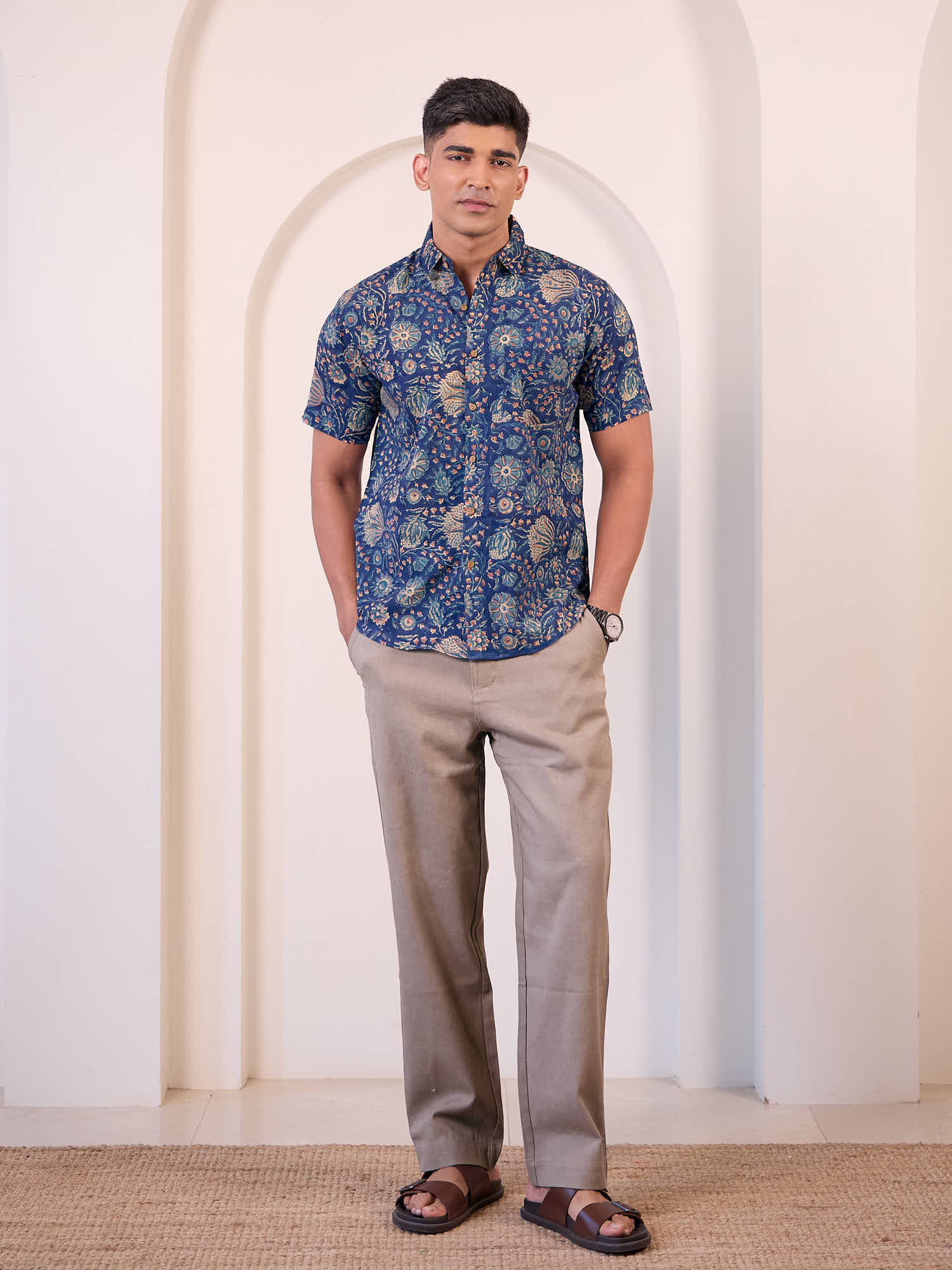 Man wearing a blue patterned shirt and beige pants standing against a white wall.