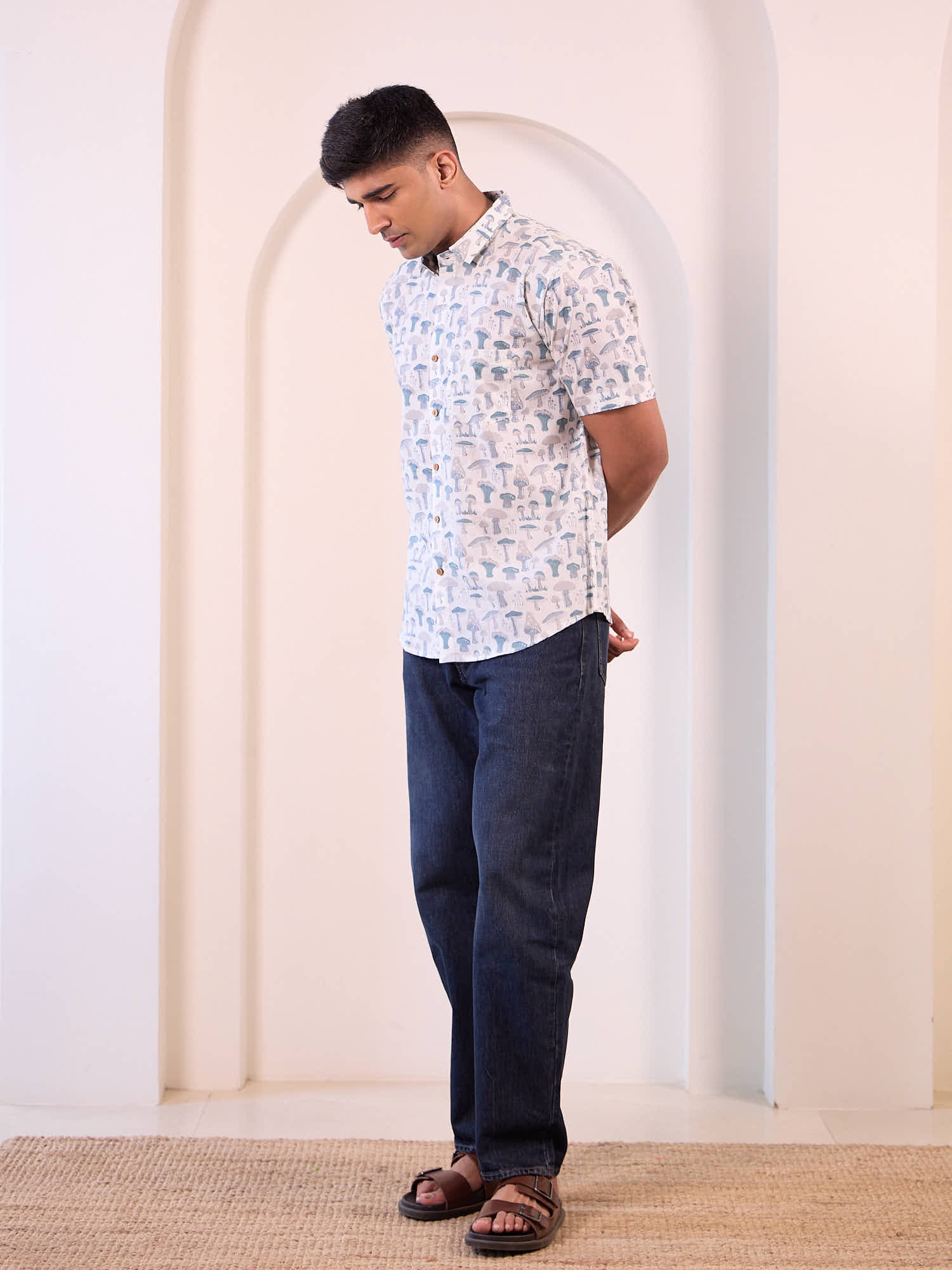 Man wearing a patterned shirt and dark jeans standing against a white wall.