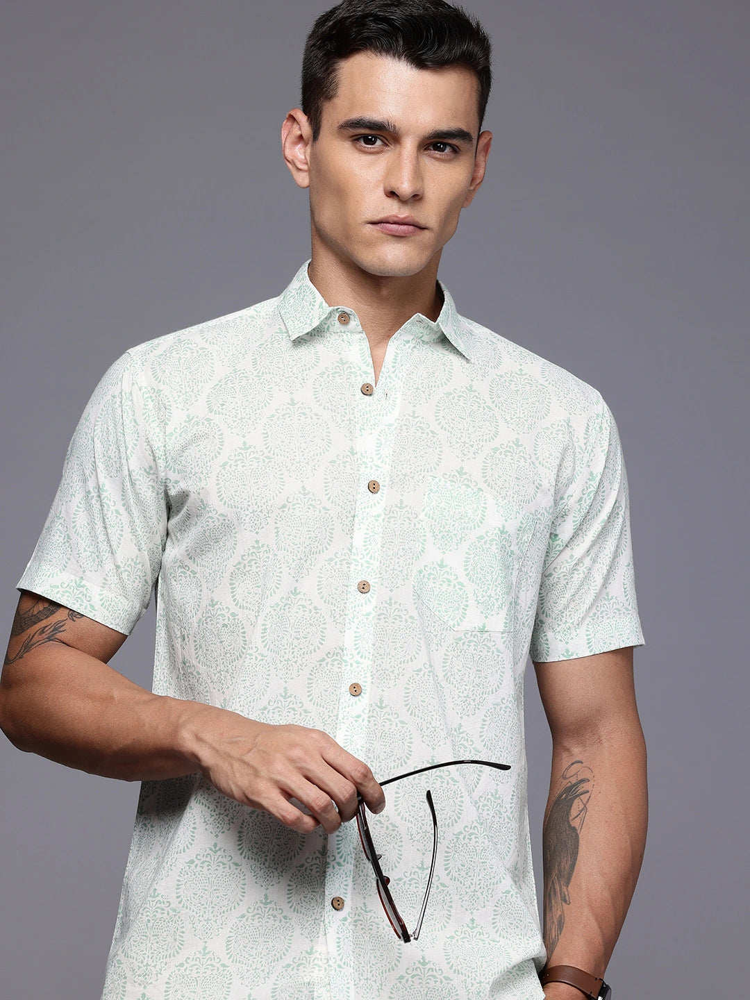 Man wearing a light green patterned shirt holding glasses against a gray background